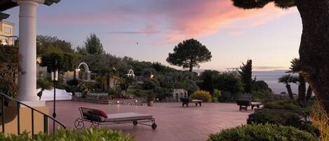 A courtyard with a sun lounger, benches, and a view of the sea at sunset.