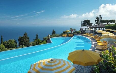 A curved infinity pool with yellow and white striped umbrellas, lounge chairs, and a view of the ocean.