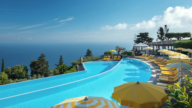 Una piscina infinita curvada con sombrillas a rayas amarillas y blancas, sillas de descanso y vista al océano.