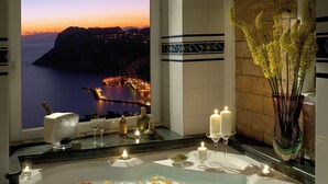Un baño con una bañera llena de pétalos de rosa y velas, un ventanal grande con vistas a la costa, y un florero con flores amarillas.