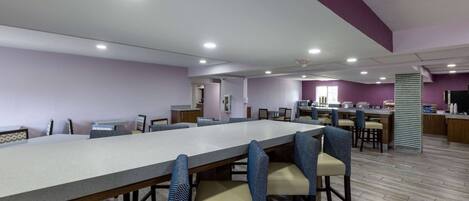 Un comedor espacioso con una larga mesa y múltiples sillas, un área de cocina al fondo, y una pared de acento color púrpura.