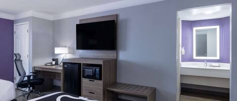A hotel room with a bed, desk, microwave, TV, and a bathroom with a mirror and sink.