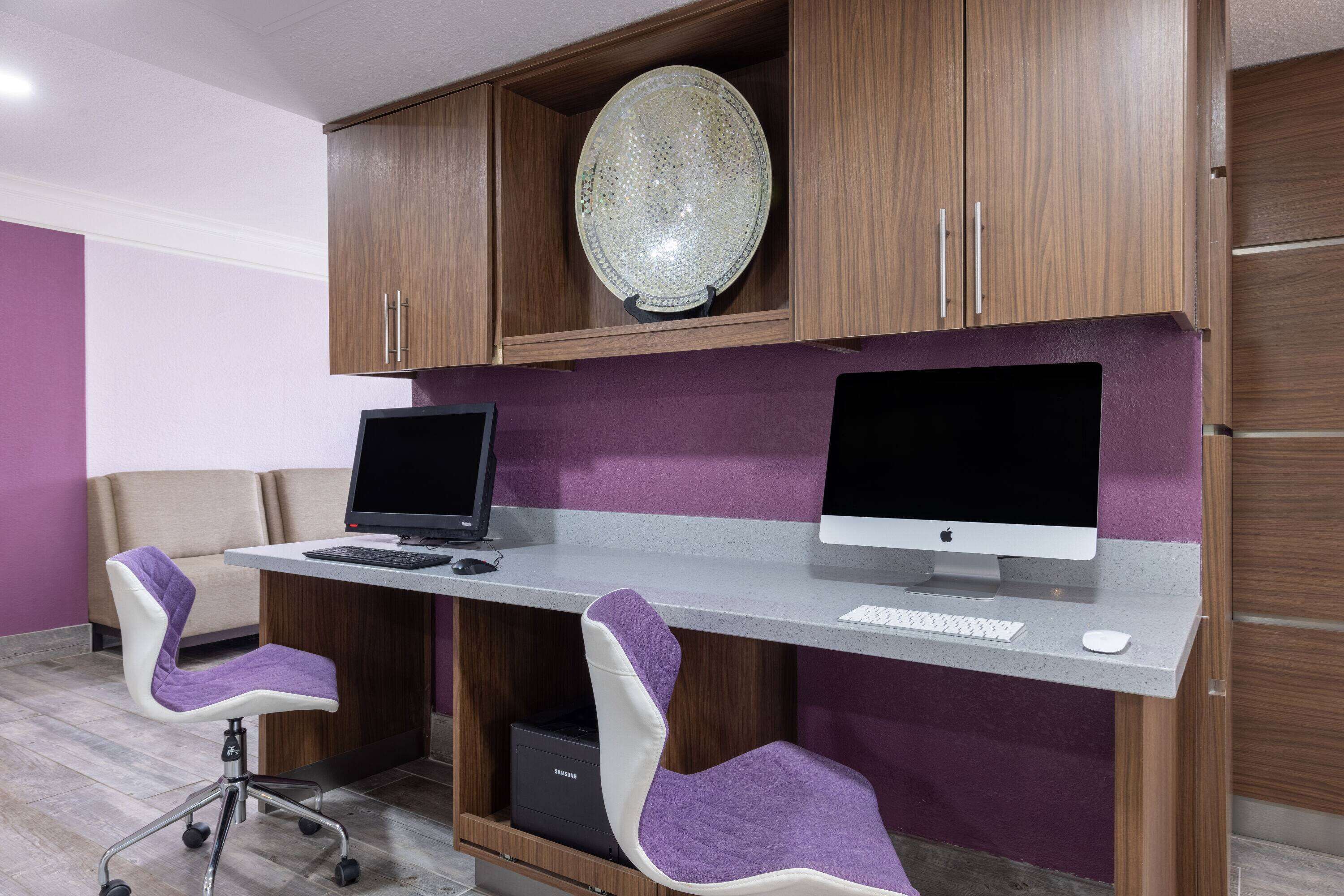 Un espacio de oficina moderno con dos escritorios para computadora, cada uno con monitor, teclado y ratón. Hay dos sillas de oficina moradas con ruedas. La habitación tiene armarios de madera y un plato decorativo en la pared.
