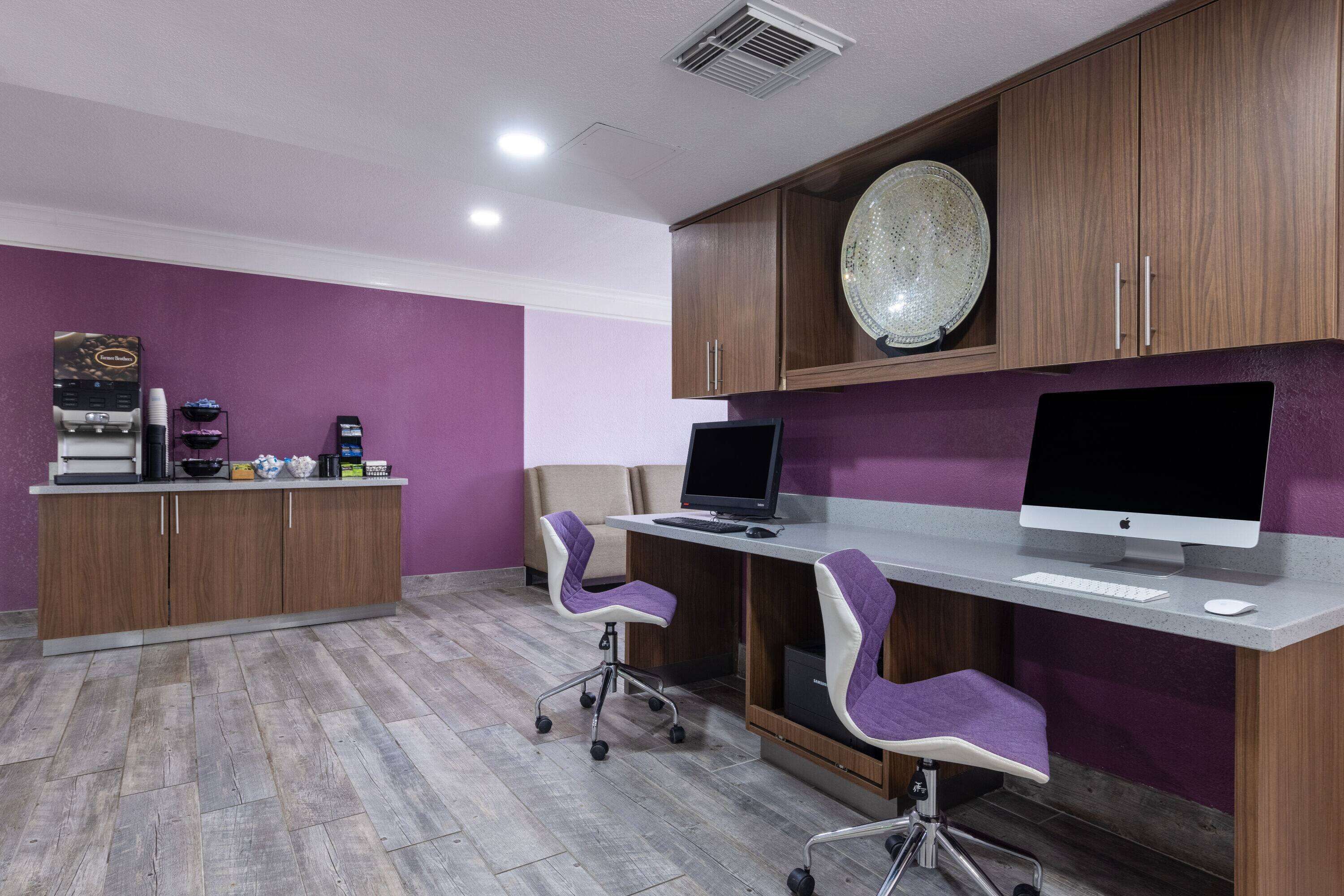 A modern office space with a purple wall, wooden cabinets, a desktop computer, two purple office chairs, and a coffee machine.
