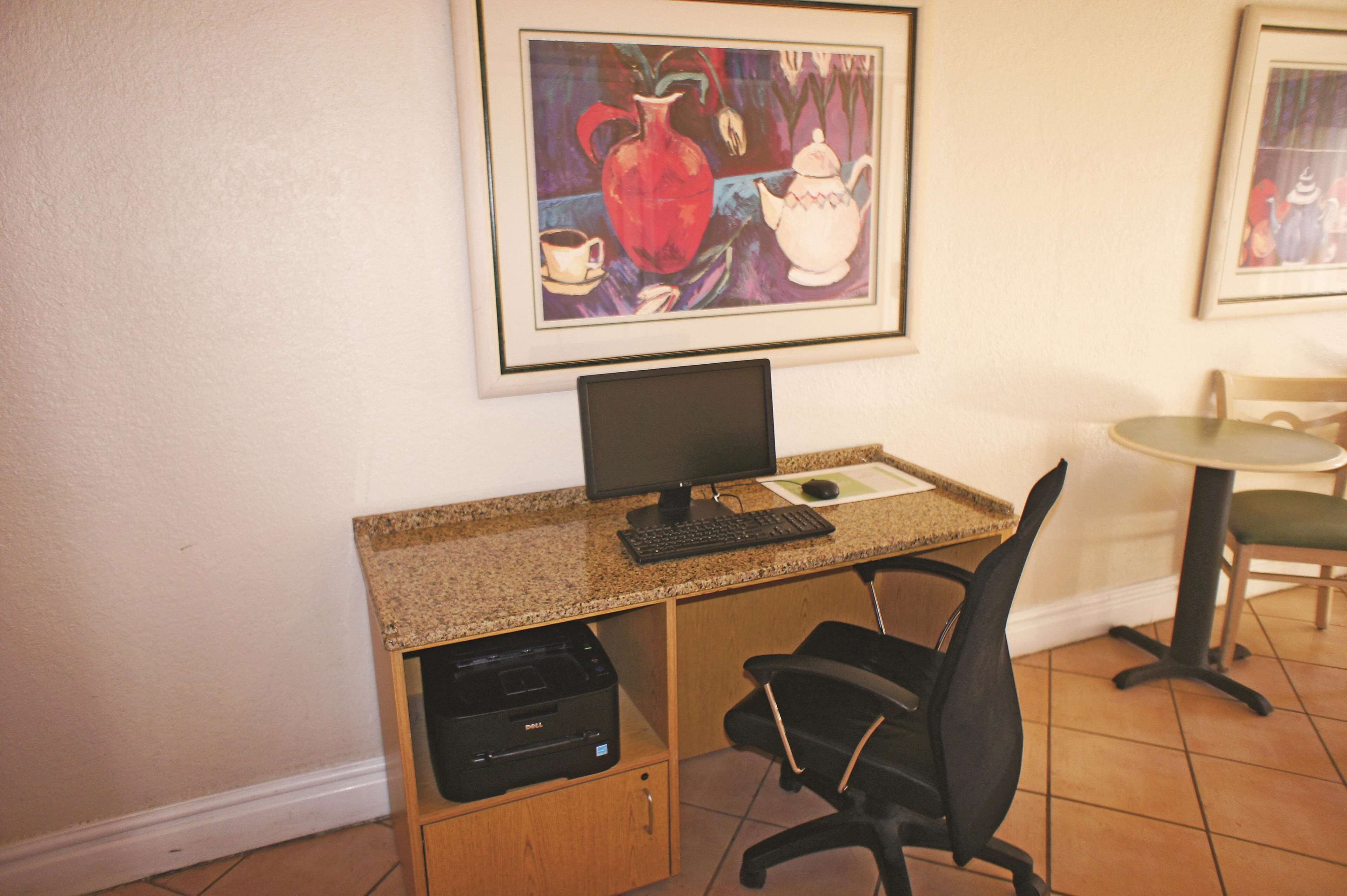 A small office space with a desktop computer, a printer, a chair, and a framed painting on the wall.