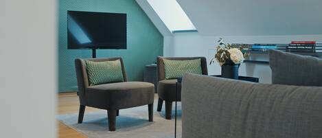 Una sala de estar moderna con un televisor de pantalla plana montado en una pared texturizada color turquesa, dos sillones oscuros con cojines estampados, un sofá gris y una estantería con libros y objetos decorativos.