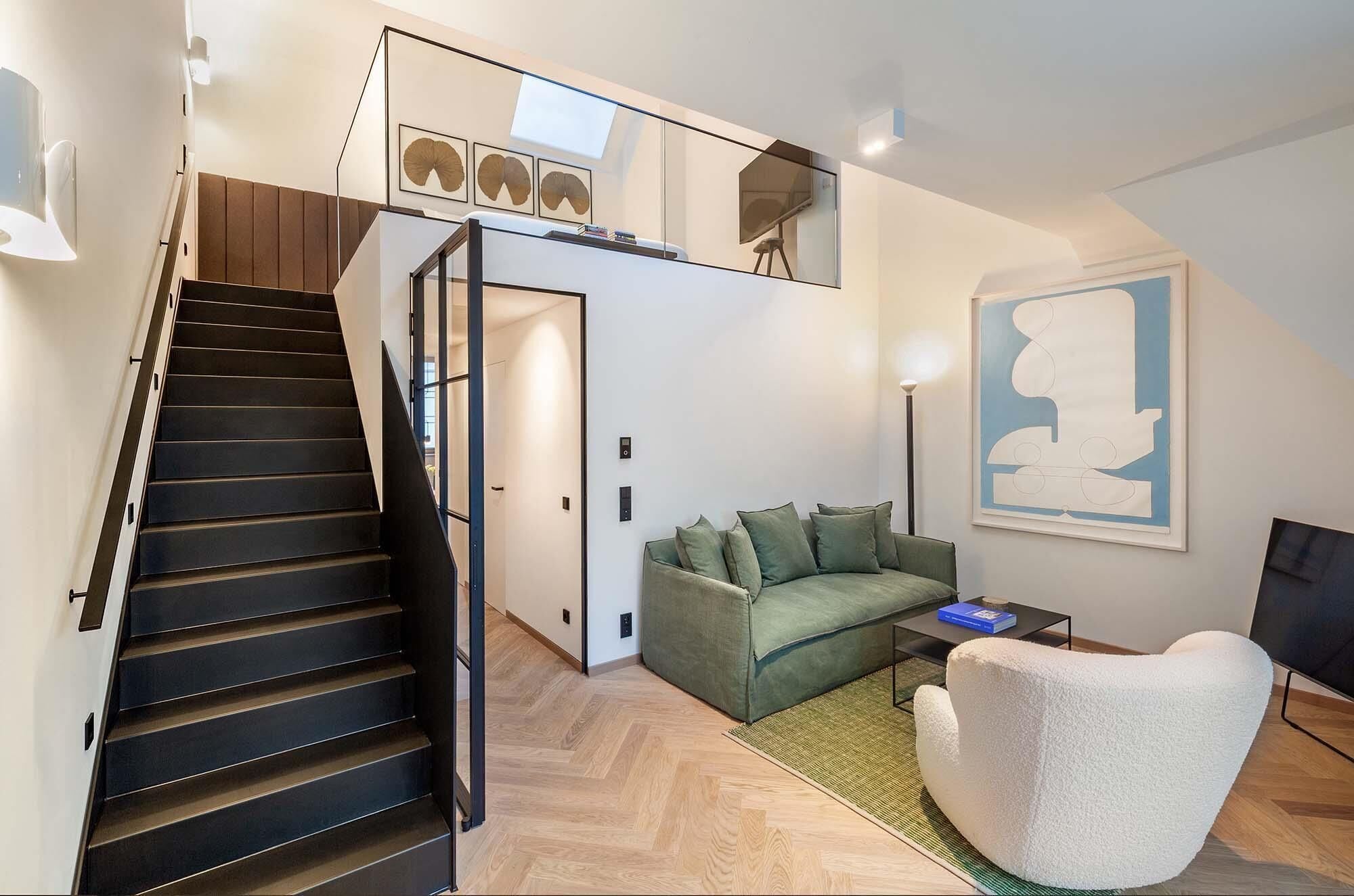 A modern interior with a staircase, a sofa, a white armchair, and a painting on the wall.