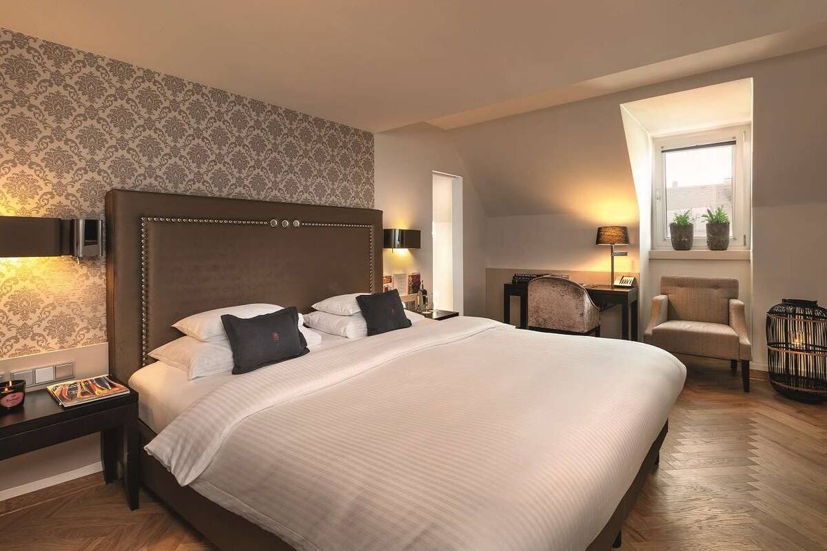 Une chambre d’hôtel avec un grand lit, une table de chevet avec une lampe, une chaise, une petite table avec une lampe et une fenêtre ornée de plantes.