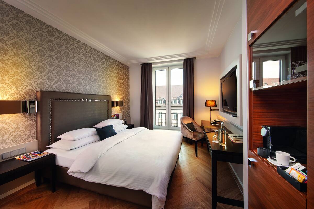 Une chambre d’hôtel avec un grand lit, un bureau avec une lampe, une chaise, une télévision et une fenêtre avec des rideaux.