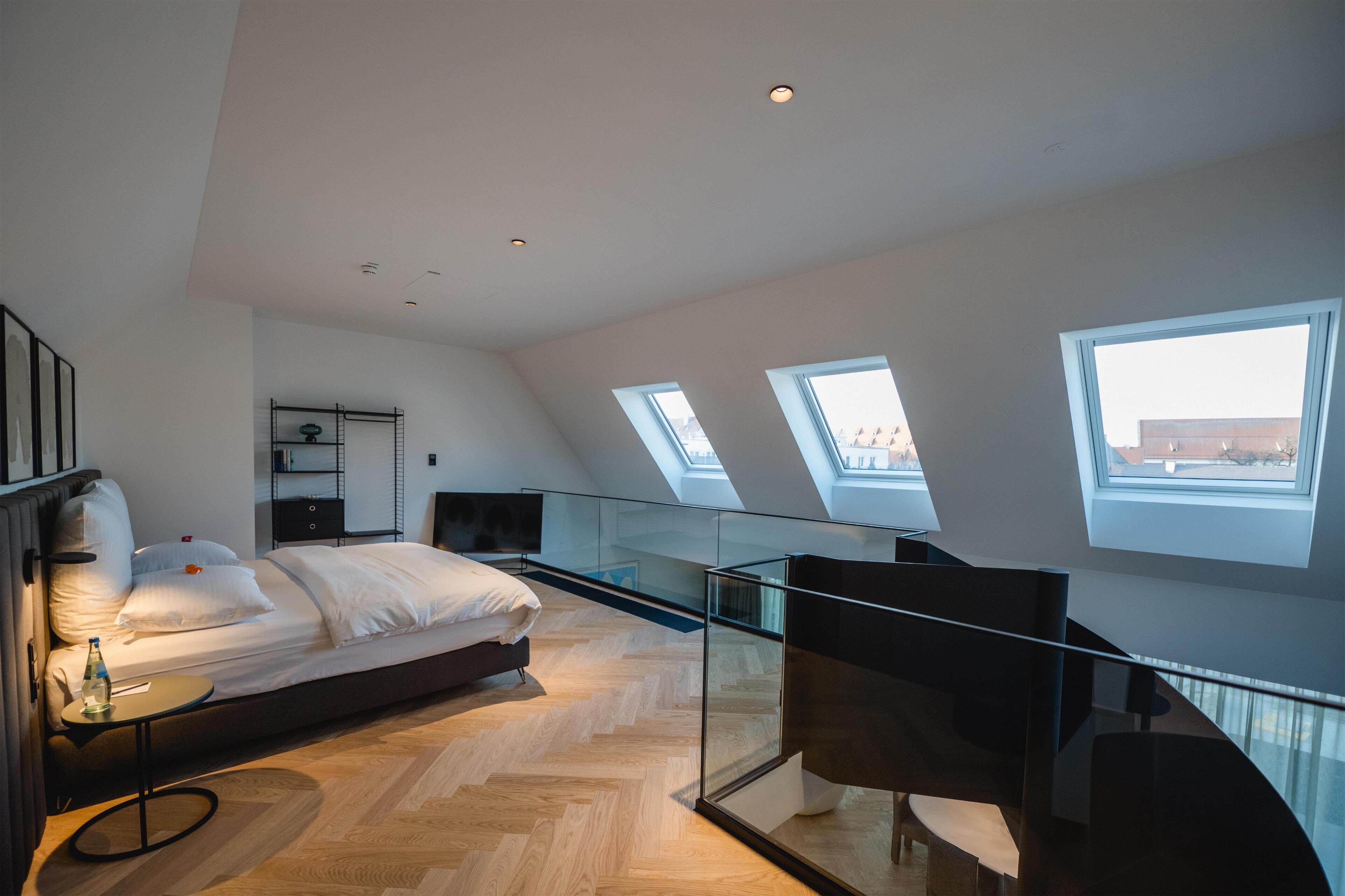 A modern bedroom with a bed, a small table, and a glass railing overlooking the city.