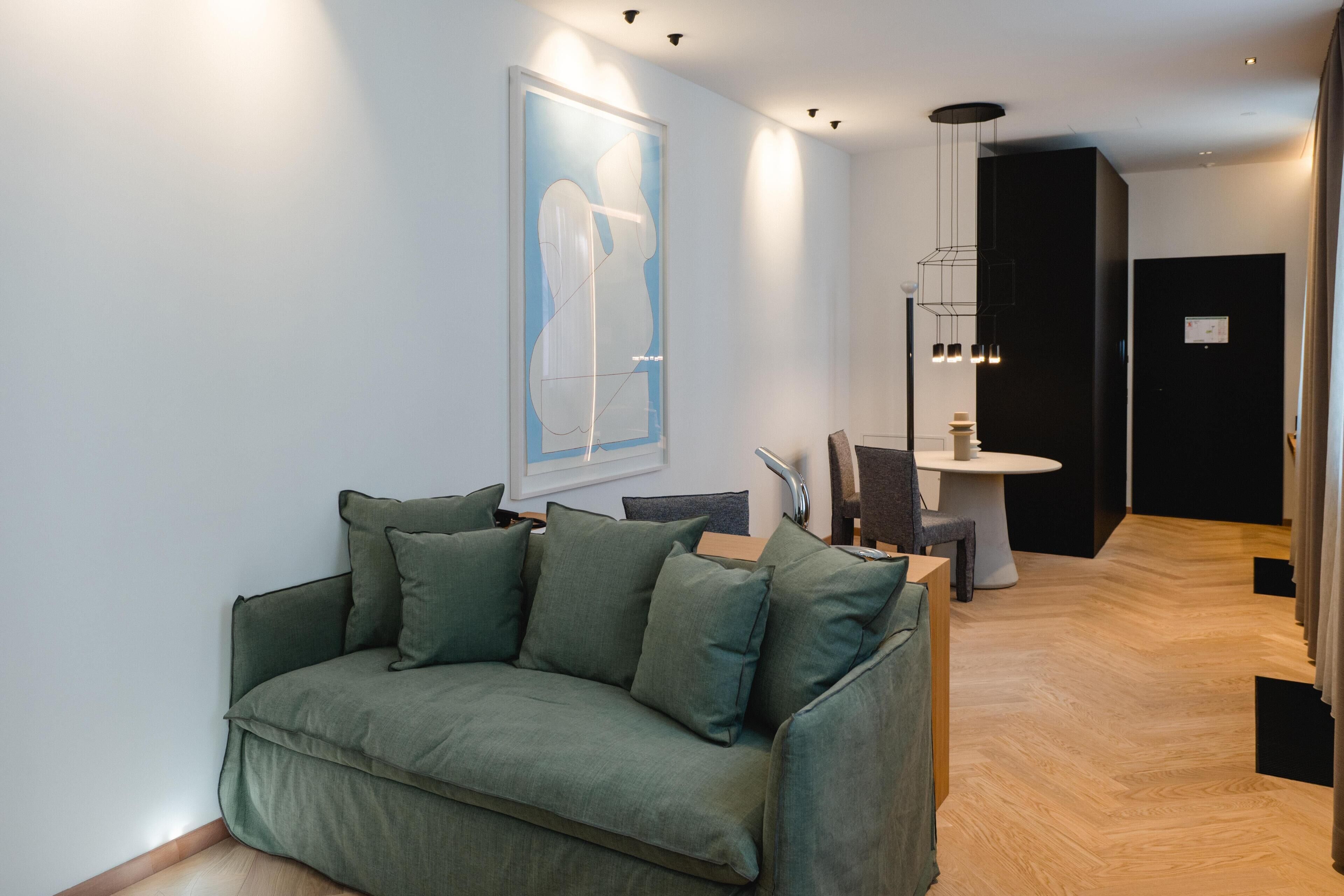 Ein modernes Wohnzimmer mit einem grünen Sofa, einem gerahmten Kunstwerk an der Wand, einem Essbereich mit Tisch und Stühlen sowie einem Flur, der in ein weiteres Zimmer führt.