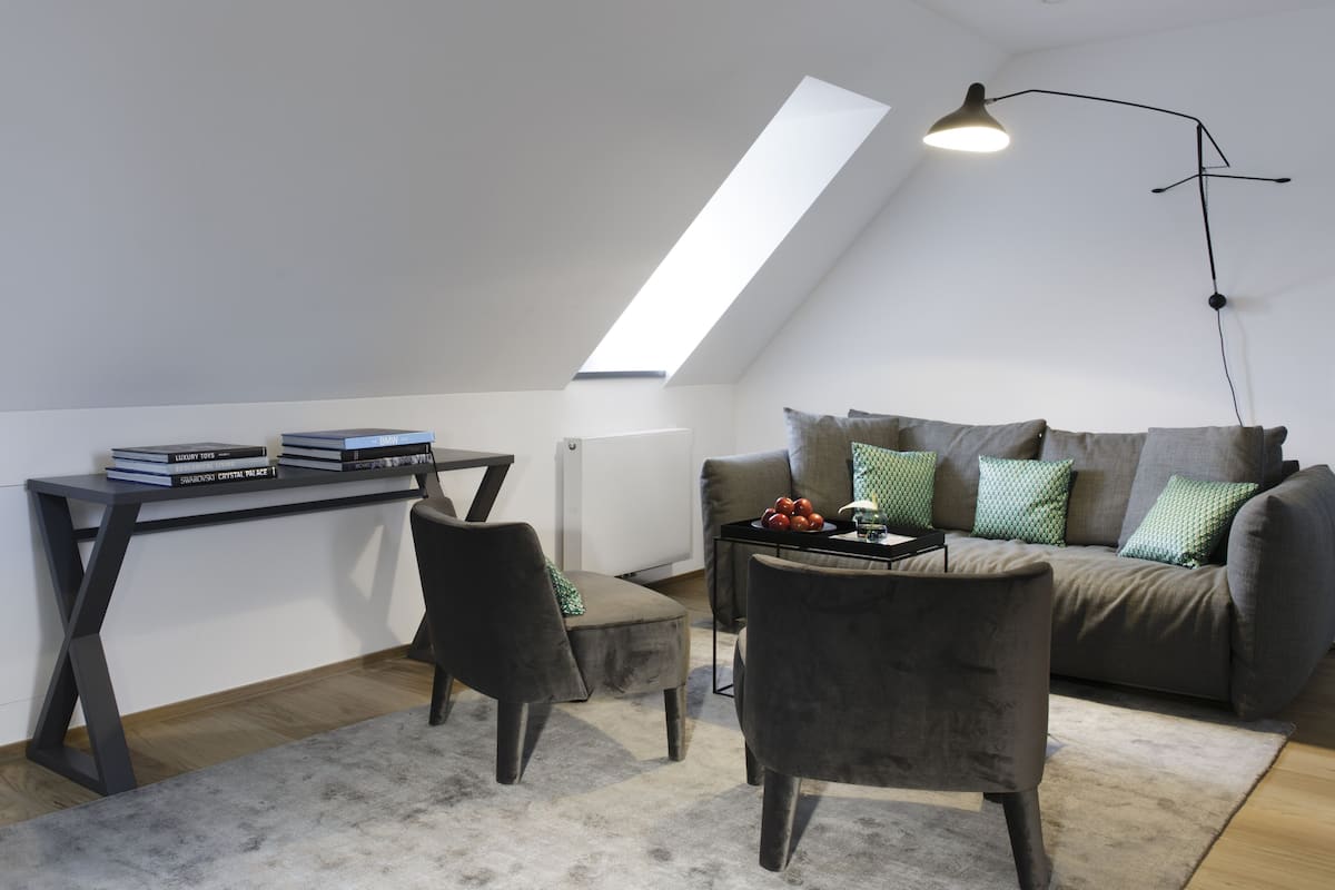 Une chambre de style loft moderne, avec un plafond en pente, une lucarne, un canapé gris agrémenté de coussins à motifs, une petite table avec des livres et un fauteuil sombre.