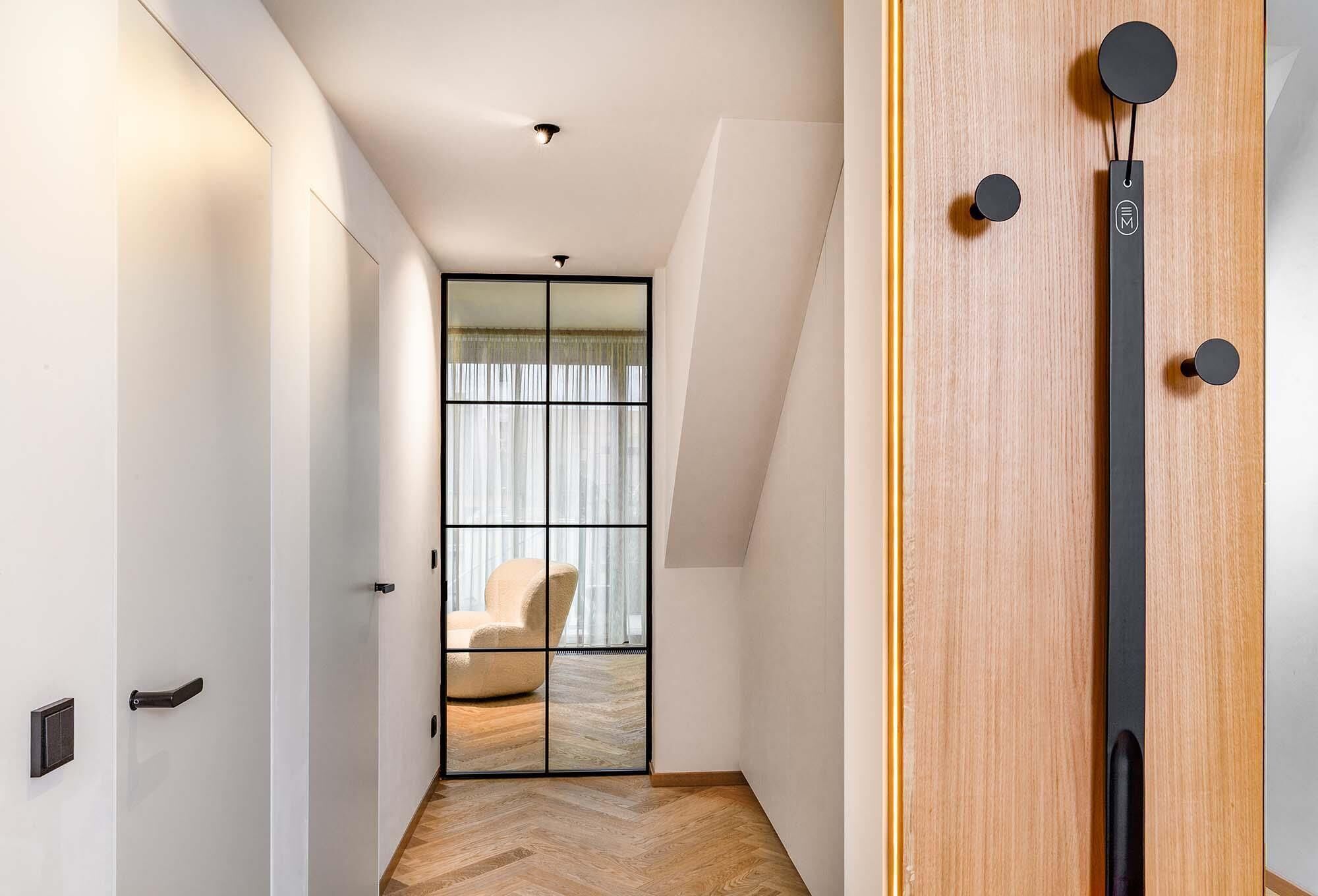Un couloir moderne avec un plancher en bois, une porte coulissante en verre donnant sur une pièce où se trouve une chaise, et une porte en bois avec une poignée noire.