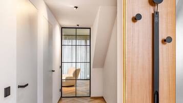 Un couloir moderne avec un plancher en bois, une porte coulissante en verre donnant sur une pièce où se trouve une chaise, et une porte en bois avec une poignée noire.