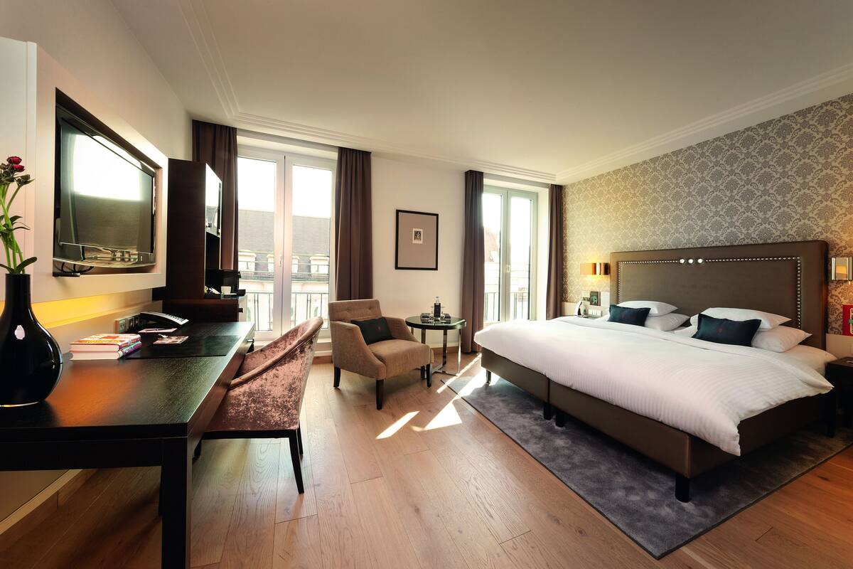 Une chambre d’hôtel avec un grand lit, un bureau avec une chaise, une petite table, un vase avec des fleurs et un téléviseur.