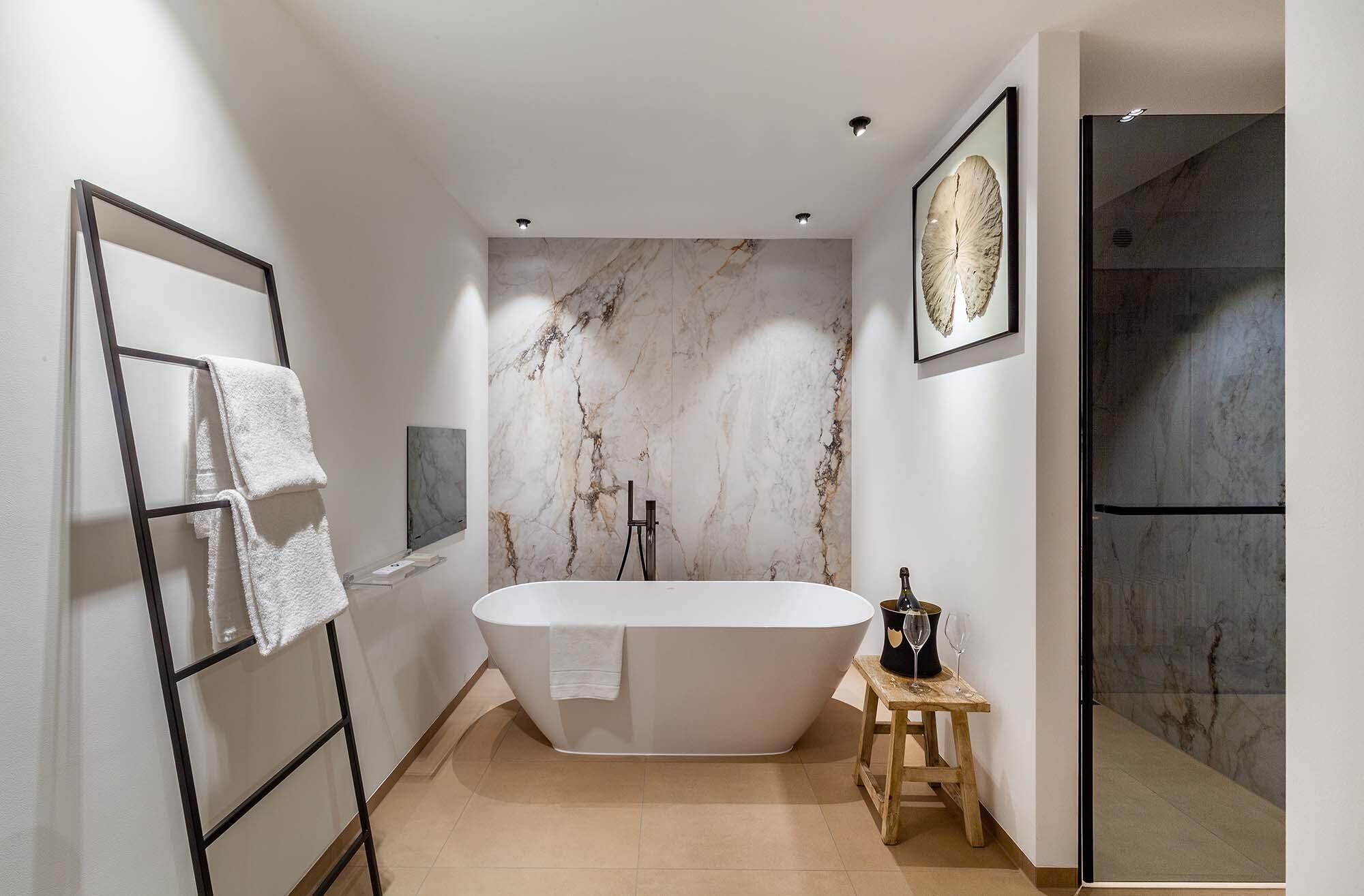 A modern bathroom with a freestanding bathtub, marble wall, and a wooden stool with a bottle and glasses.
