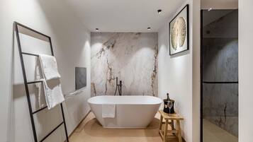 A modern bathroom with a freestanding bathtub, marble wall, and a wooden stool with a bottle and glasses.