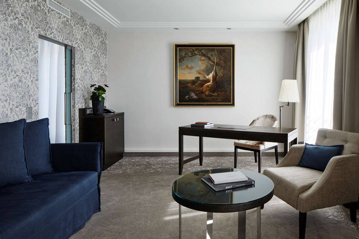 Une chambre d’hôtel avec un canapé bleu foncé, un fauteuil beige, une table basse en verre sur laquelle repose un livre, un tableau accroché au mur et un bureau avec une lampe.