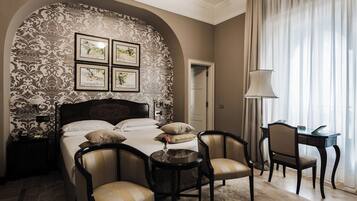 A bedroom with a large bed, two armchairs, a small table, and a desk. There are framed pictures on the wall above the bed.