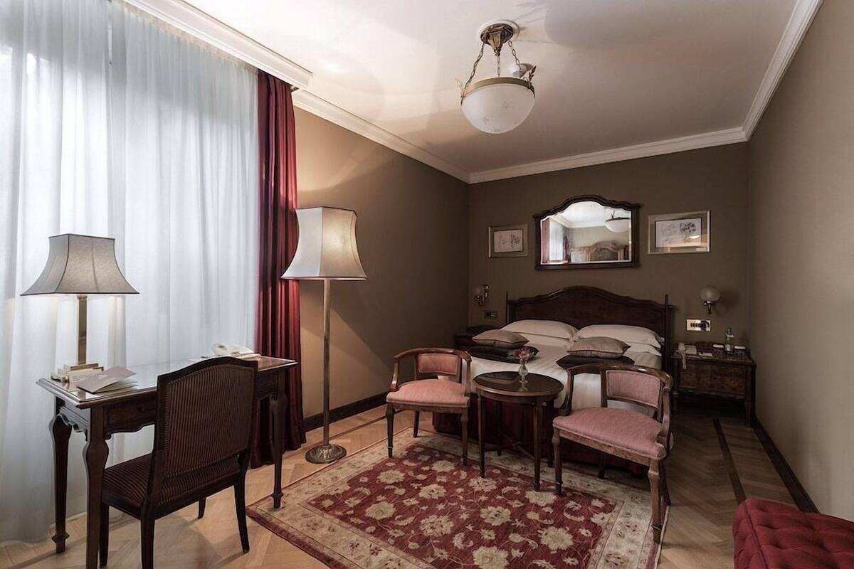 A hotel room with a bed, bedside tables, a desk with a lamp, a chair, a small round table, and a red patterned rug.