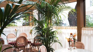 Un espace de restauration intérieur avec des chaises et des tables en osier, de grandes plantes en pot et un plafond en verre qui laisse entrer la lumière naturelle.