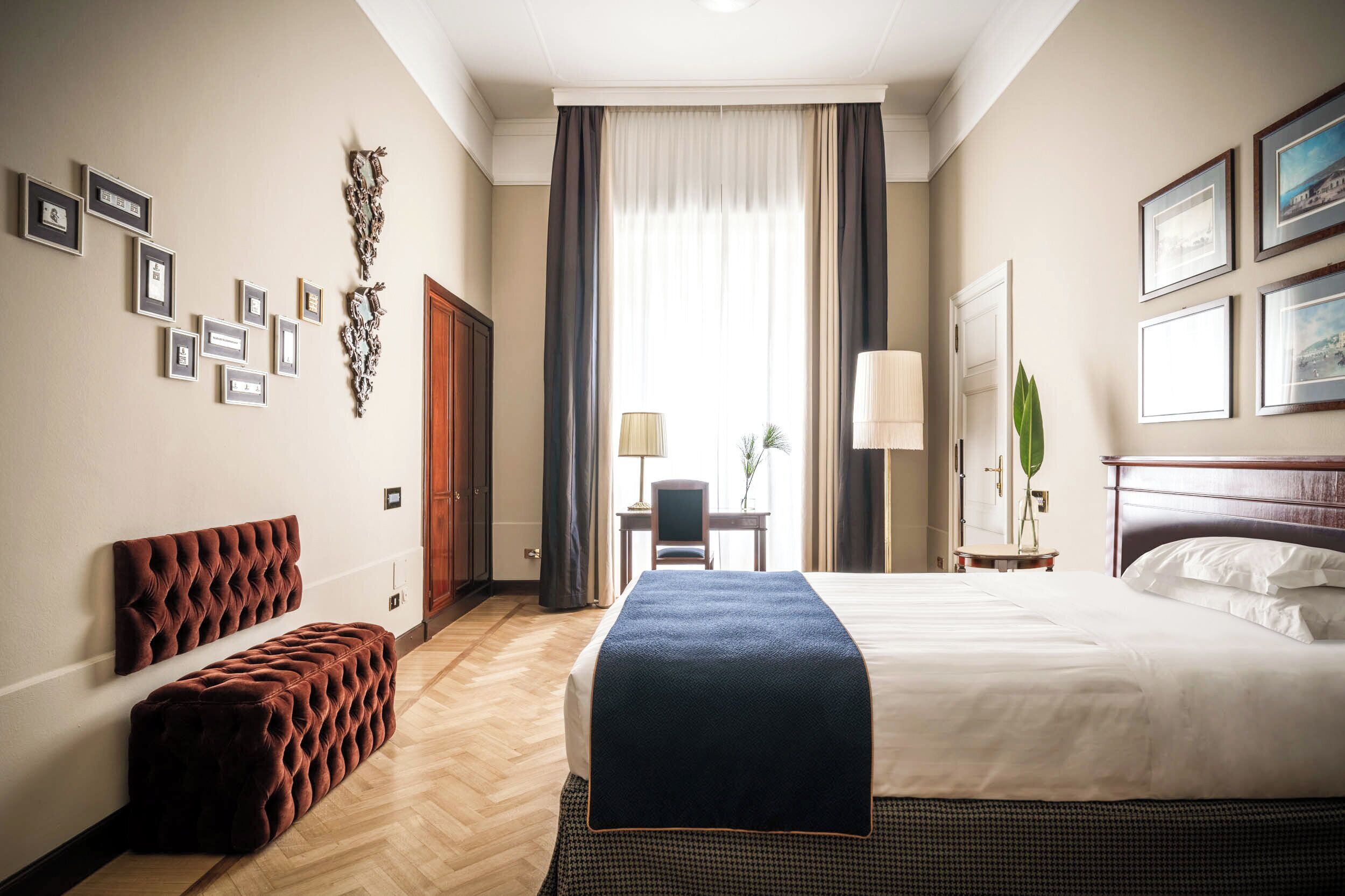 Une chambre d’hôtel avec un grand lit, un banc en bois, un bureau avec une lampe et des tableaux encadrés aux murs.