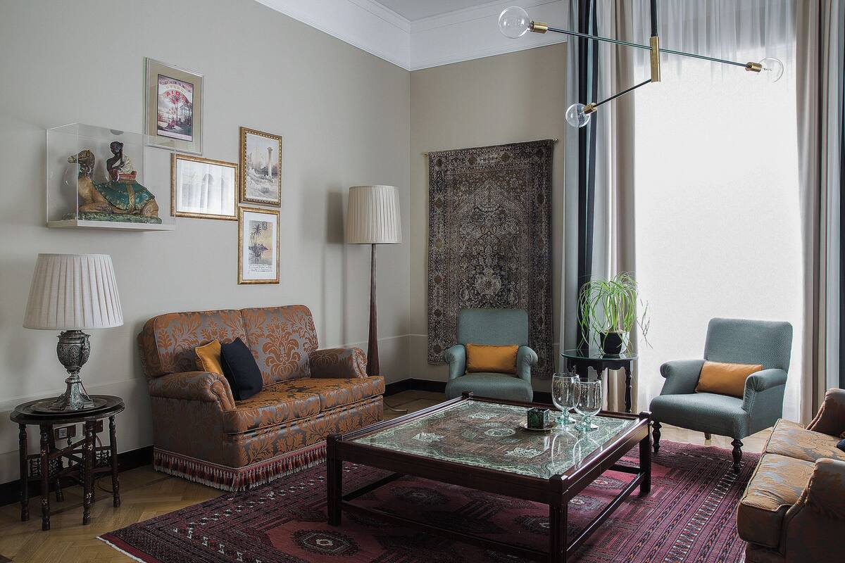 A living room with a sofa, armchairs, a coffee table, and a large rug. There are framed pictures on the wall and a lamp on a side table.