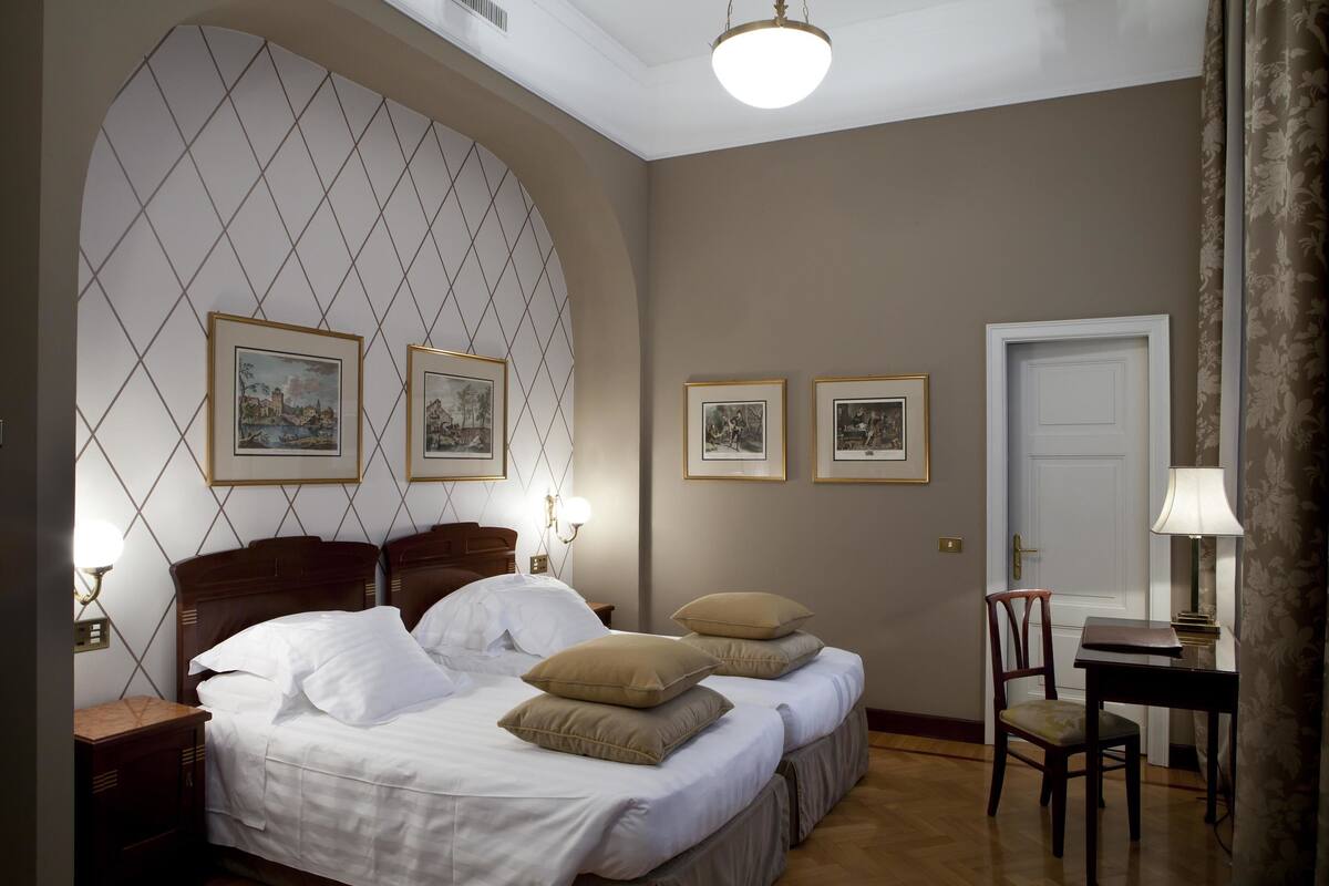 Une chambre d’hôtel avec un lit, deux tables de chevet, une chaise, un bureau et des tableaux encadrés accrochés au mur.