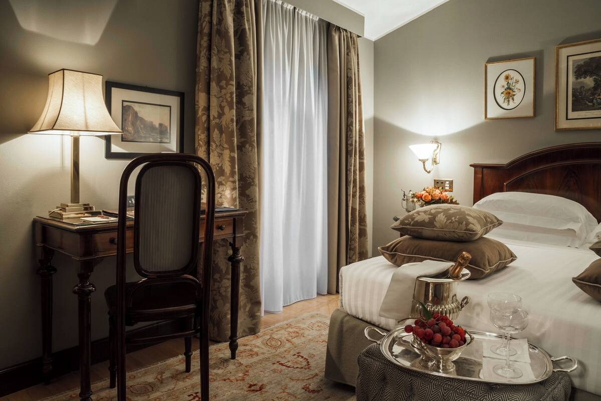 A hotel room with a bed, a desk, a chair, a lamp, a bedside table with a tray of fruit and a wine glass, and curtains.