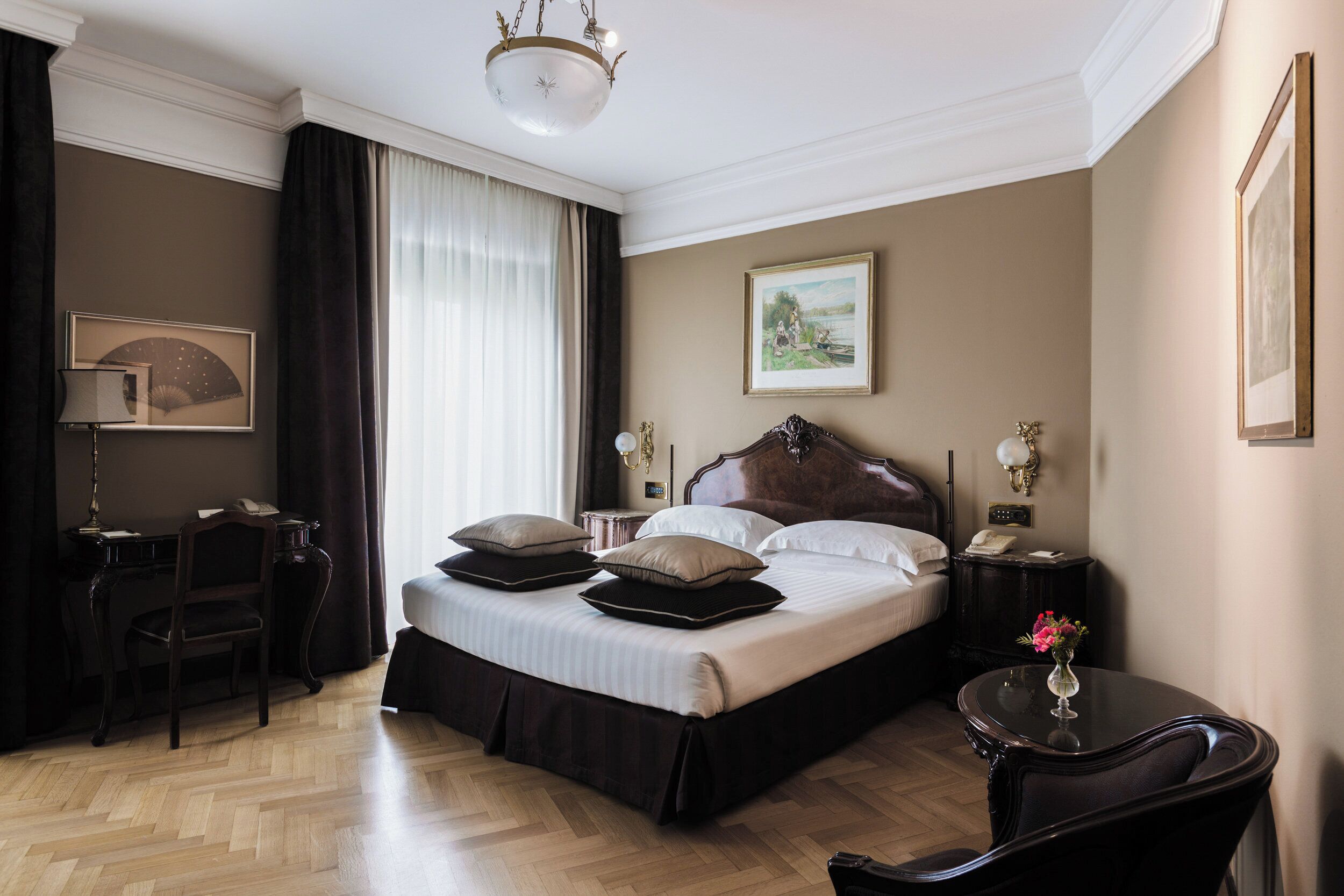 Una camera d'albergo con un letto grande, una scrivania, una sedia, un comodino e un vaso con fiori.