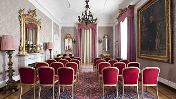 Una sala formale con sedie rosse disposte in file, un tappeto decorato e arredi sfarzosi tra cui un lampadario e opere d'arte incorniciate.