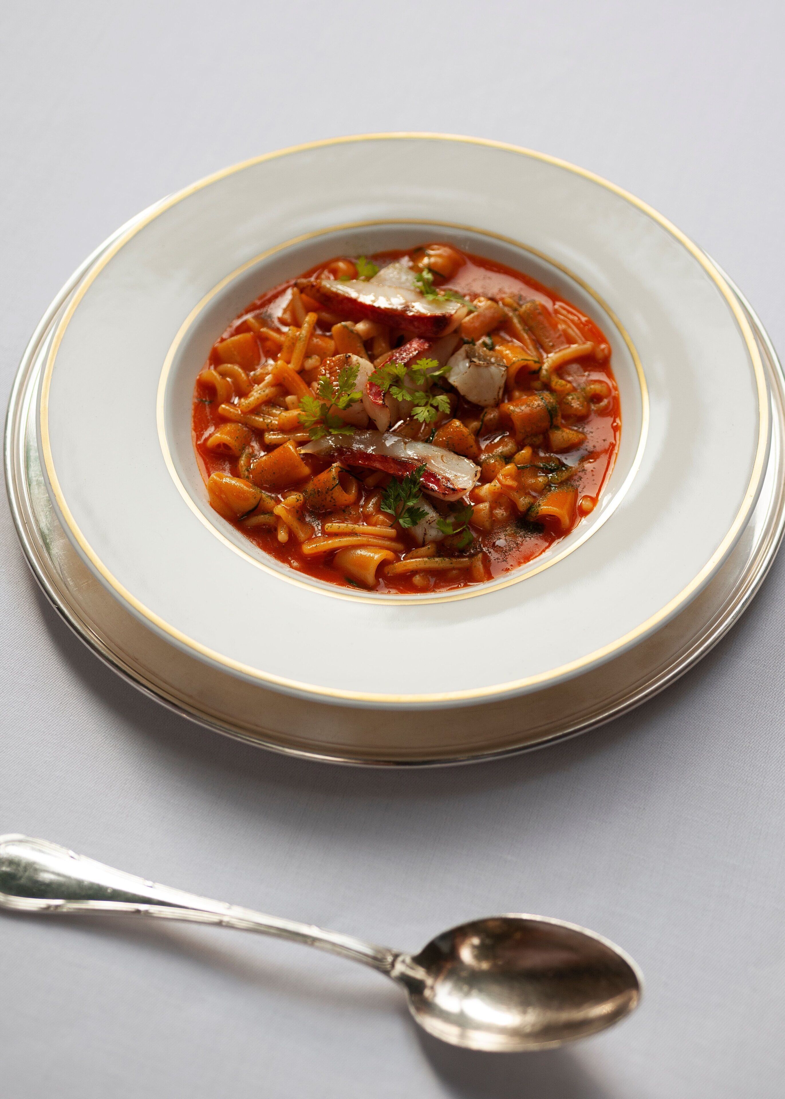 A bowl of pasta soup with a spoon beside it.