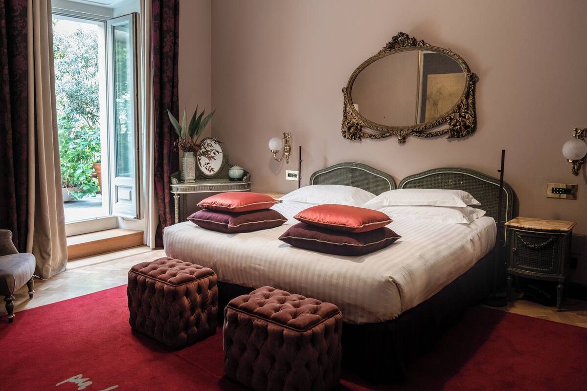A bedroom with a large bed, two ottomans, a mirror, and a nightstand.
