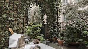 Une table dressée avec une bouteille de champagne, des verres et un plateau de desserts, dans un espace extérieur aménagé en jardin, avec des plantes en pot et une fontaine.