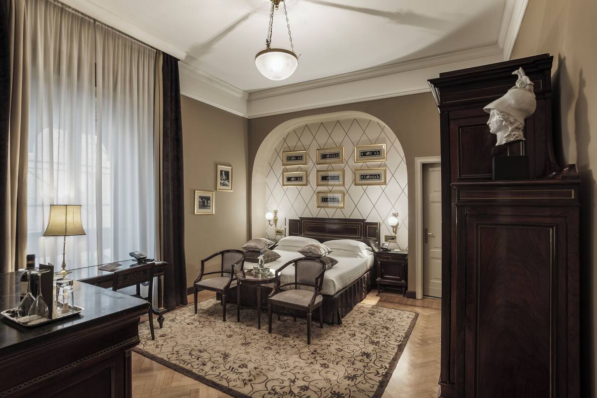 Une chambre d’hôtel comprenant un lit, des tables de chevet, un bureau avec une lampe, une chaise, un tapis et un mur décoratif orné de tableaux.