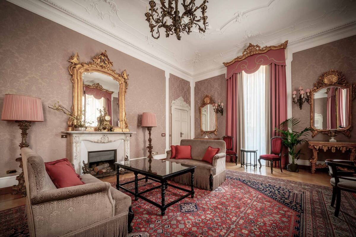 A room with a fireplace, two armchairs, a coffee table, and a large mirror with ornate gold detailing.