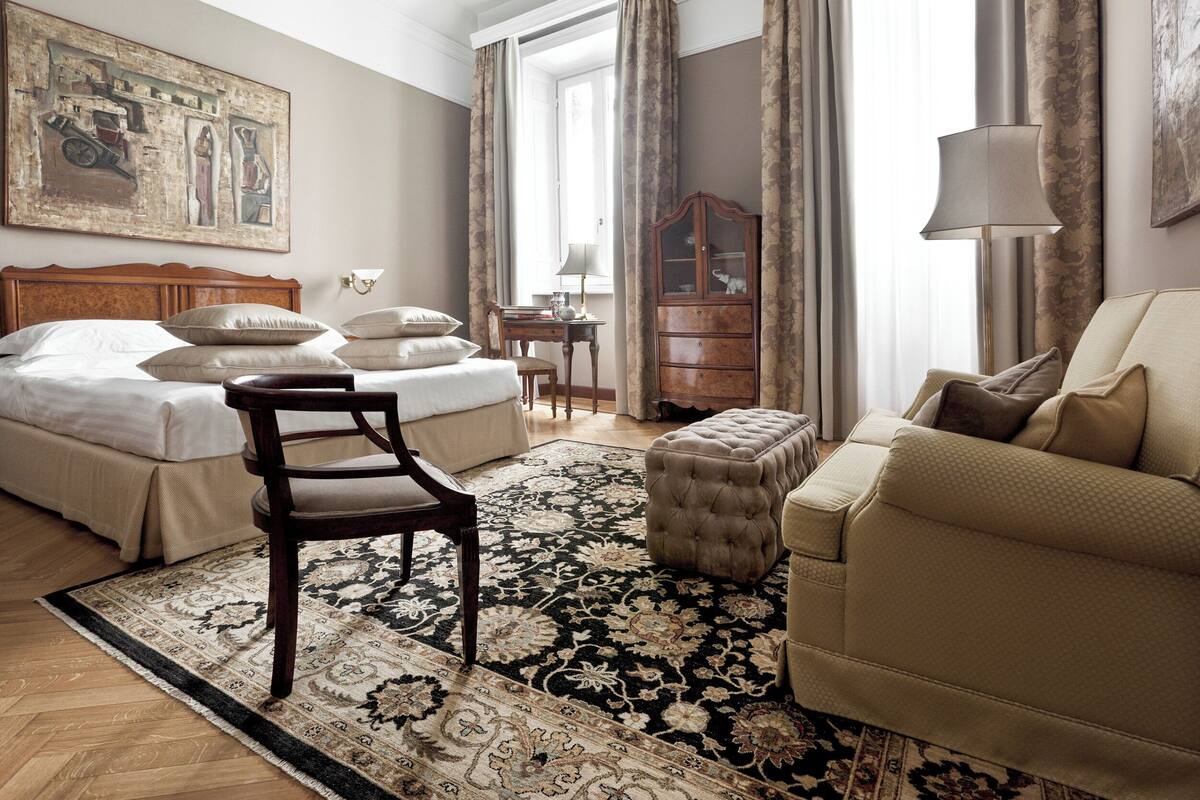 A hotel room with a bed, a chair, a sofa, a lamp, a cabinet, and a large window with curtains.