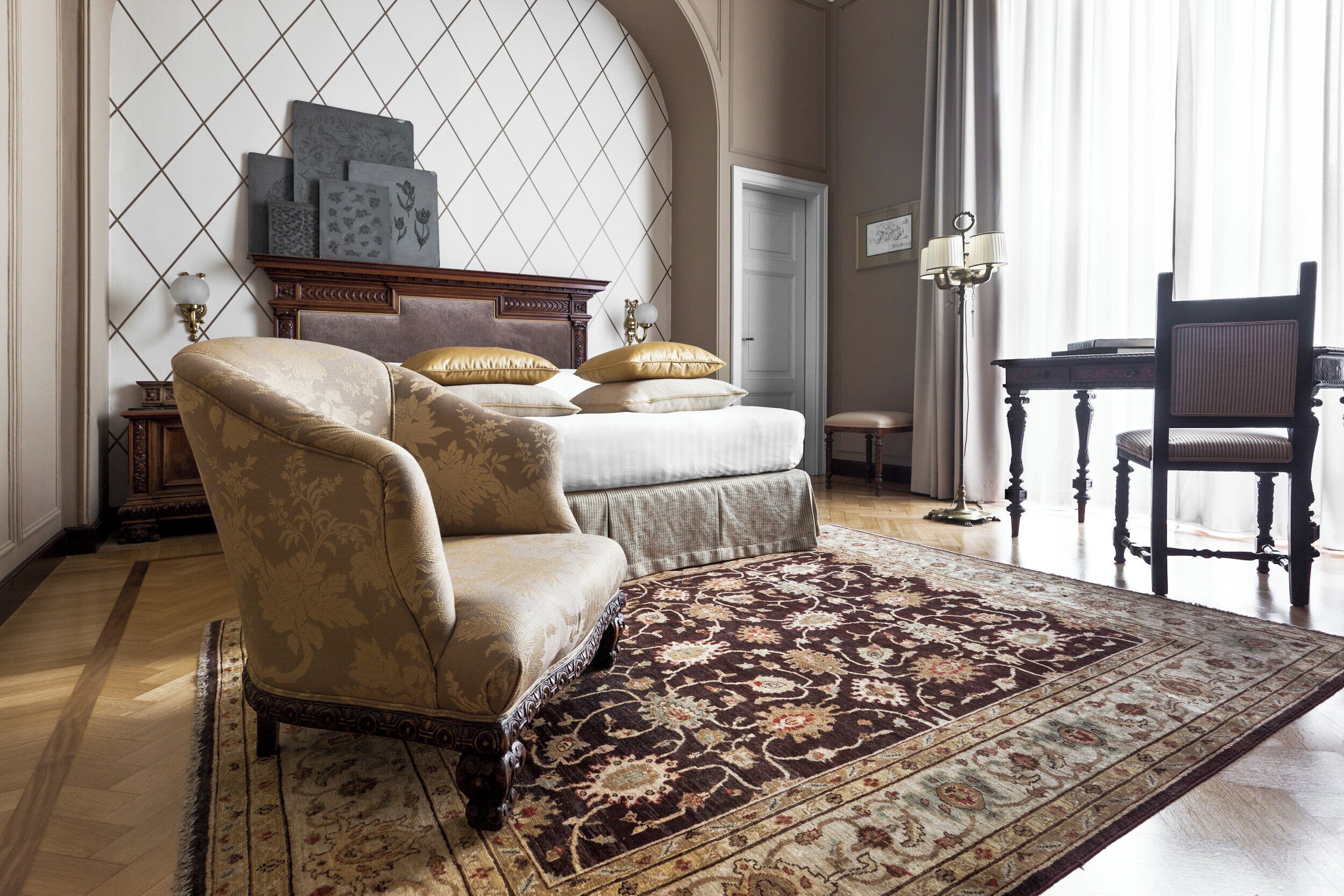 A bedroom with a large bed, two armchairs, a wooden desk, and a patterned rug.