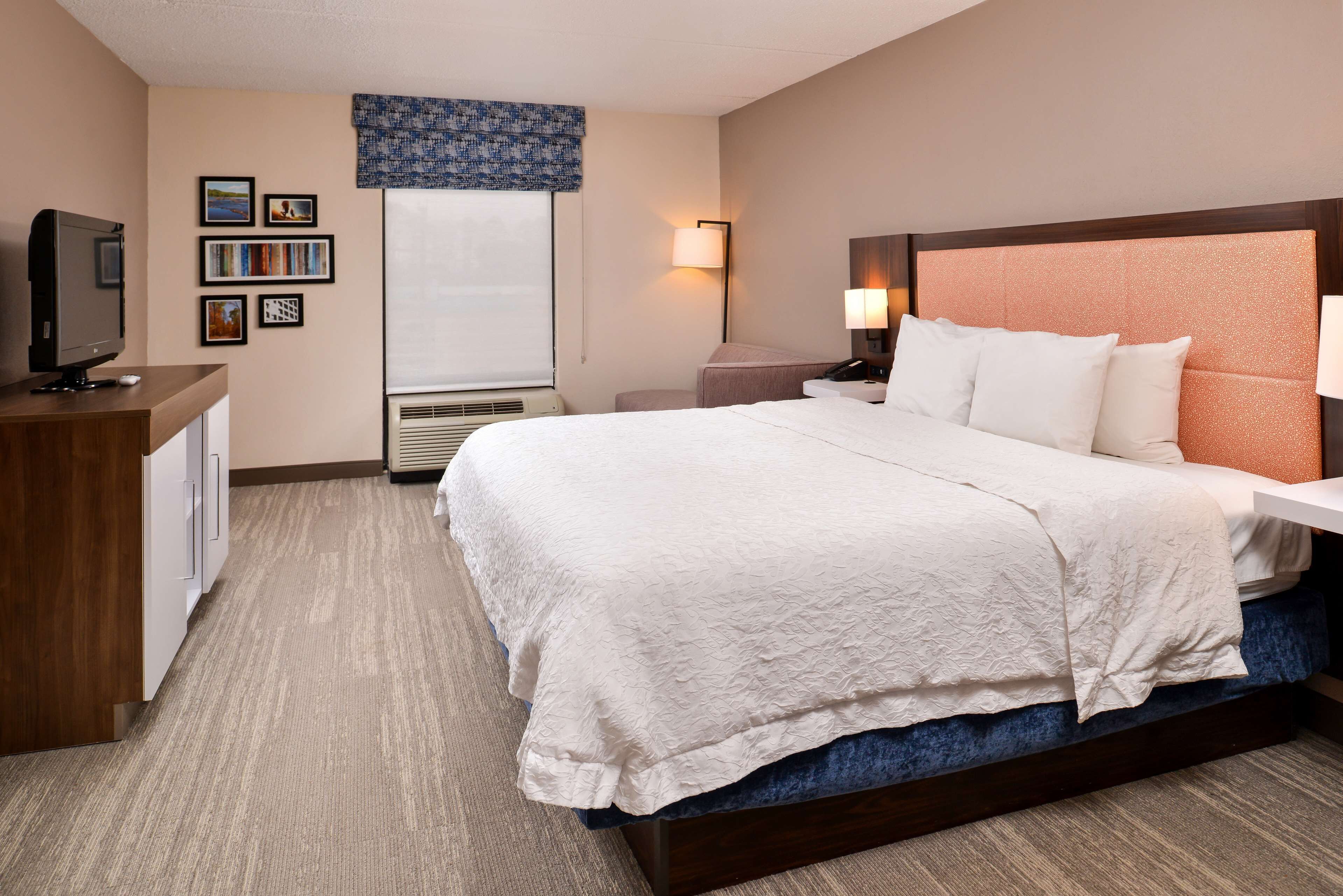 A hotel room with a large bed, a desk with a television, a wall-mounted lamp, and framed pictures.