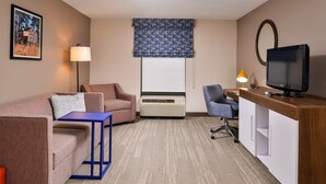 A hotel room with a sofa, a small table, a desk with a TV, a lamp, and a large window with a blue curtain. - Hampton Inn Emporia (Emporia)