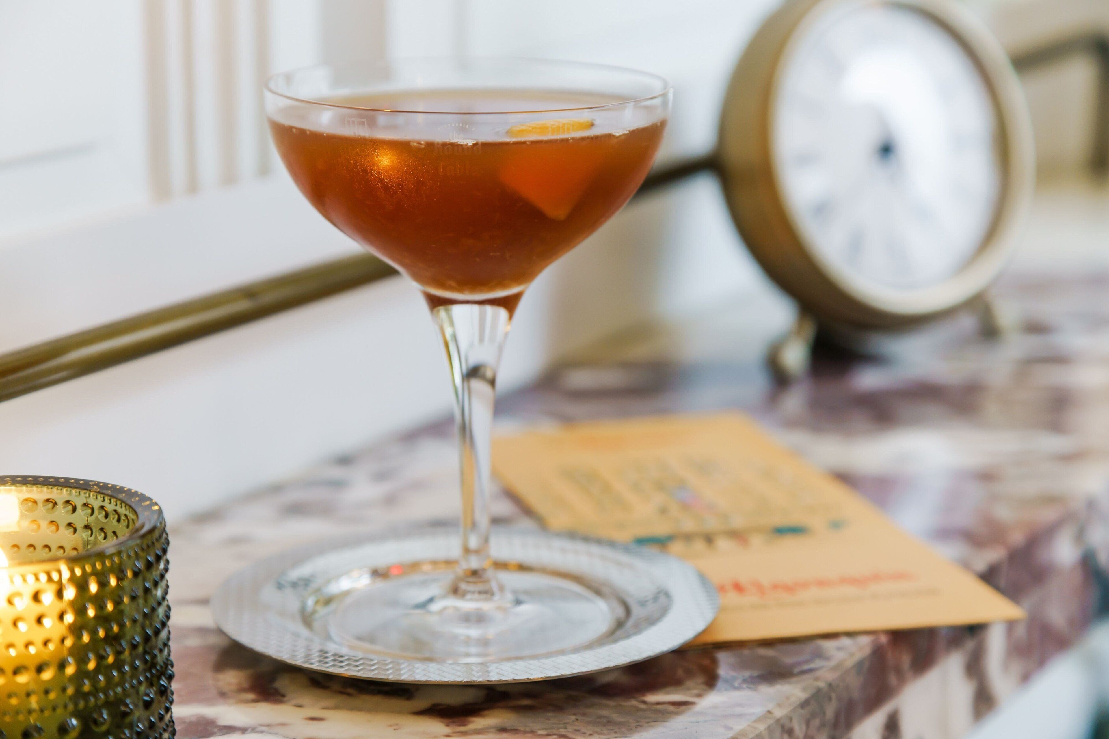 Een martini glas op een marmeren tafel, met een klok en een menukaart op de achtergrond.