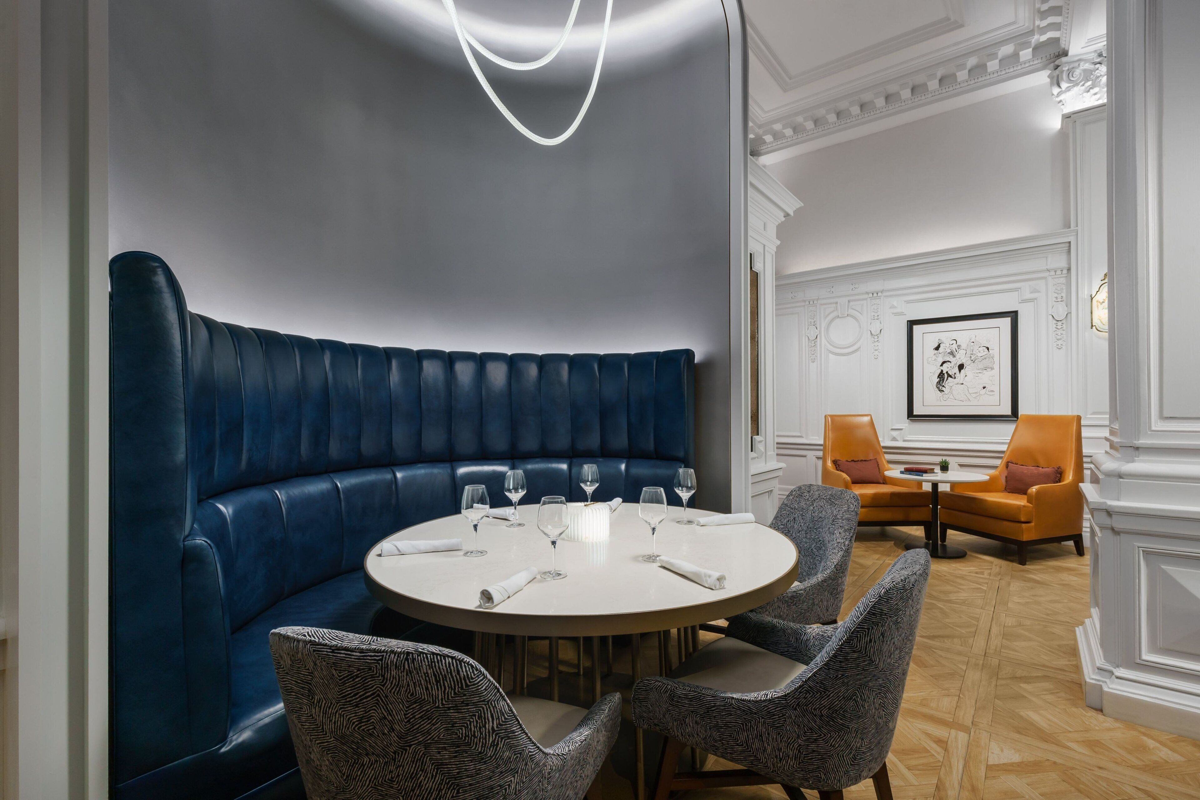 Una mesa de comedor redonda con seis sillas, un banco tapizado azul y una pared blanca con un cuadro enmarcado.
