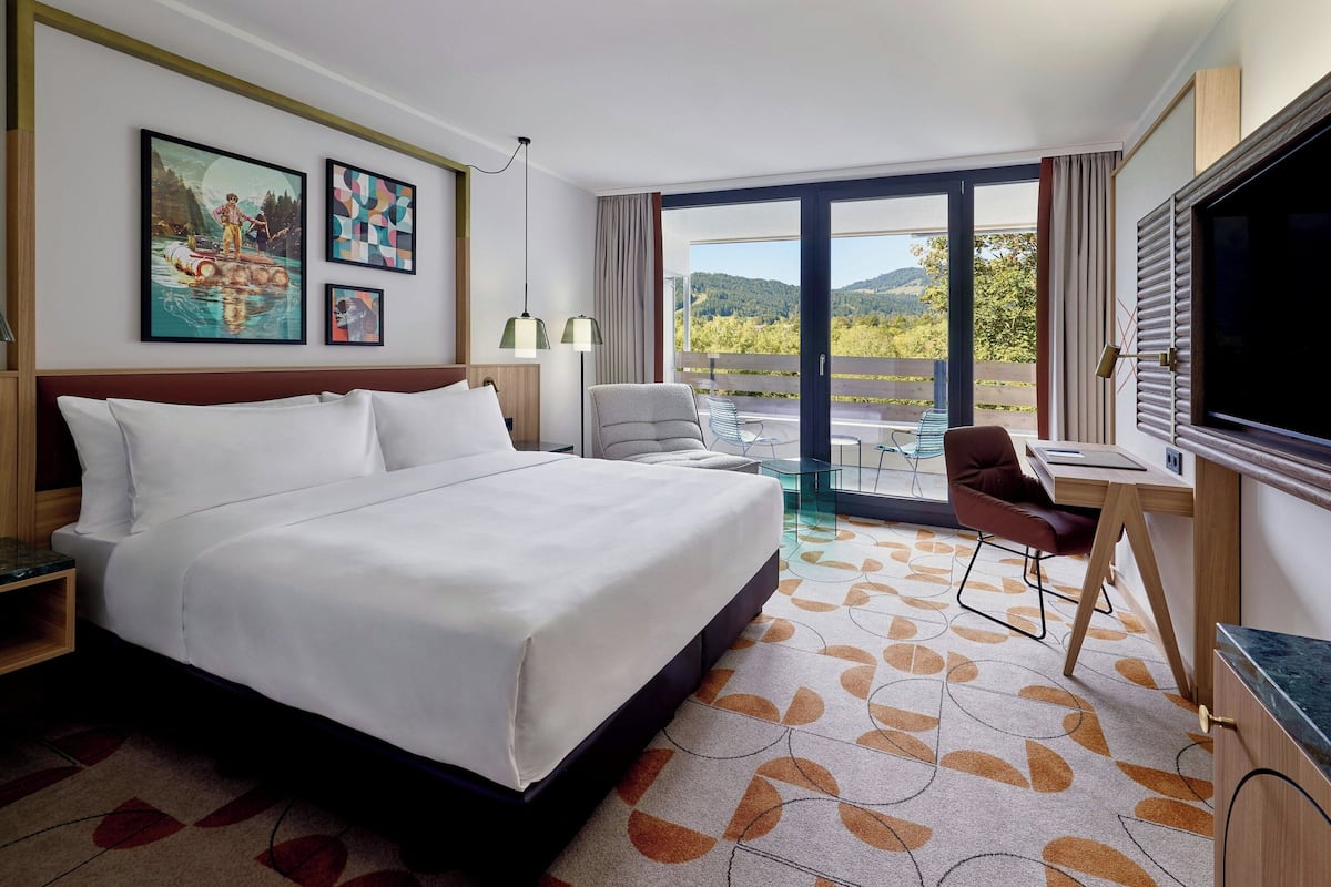 A hotel room with a large bed, a desk with a chair, a TV, and a balcony with a view of the mountains.