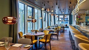 A modern restaurant with wooden tables and yellow chairs, large windows, and hanging pendant lights. - Flesslers Lenggries, a Tribute Portfolio Hotel (Lenggries)