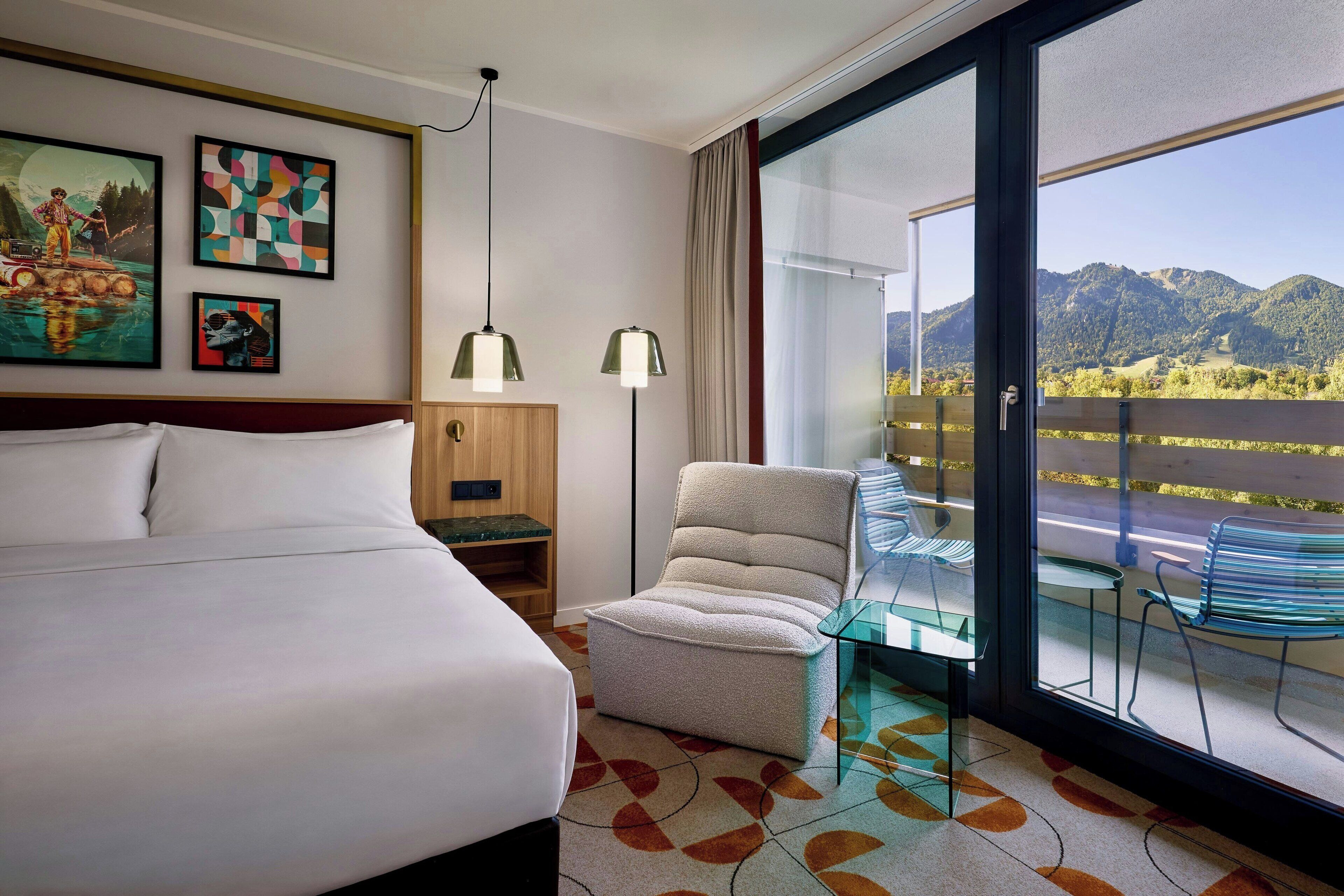 A hotel room with a large bed, a small armchair, a glass table, and a balcony with a view of mountains.