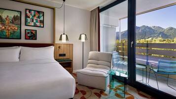 Habitación de hotel con una cama grande, un sillón pequeño, una mesita de cristal y un balcón con vistas a las montañas.