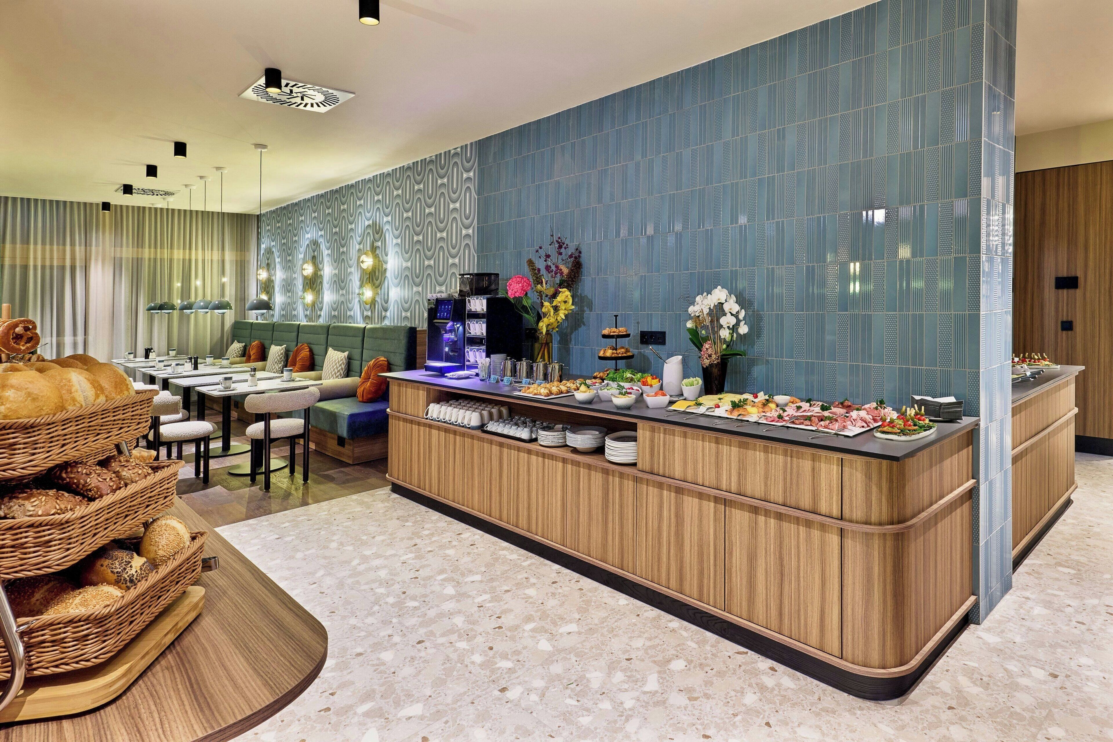 Un moderno vestíbulo de hotel con un área de bufé que ofrece diversas opciones de comida, una máquina de café y un espacio de estar con sofás y mesas.