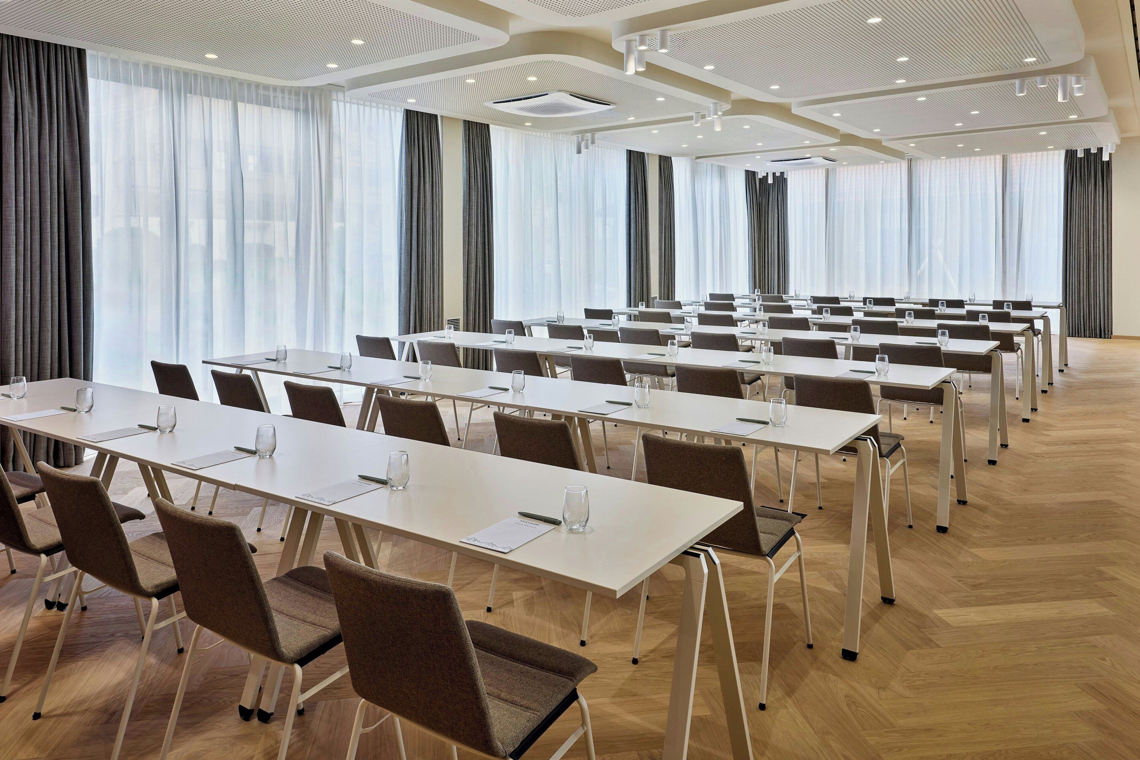Ein Konferenzraum mit mehreren Tischen und Stühlen, großen Fenstern mit Vorhängen und einem modernen Deckenstil.