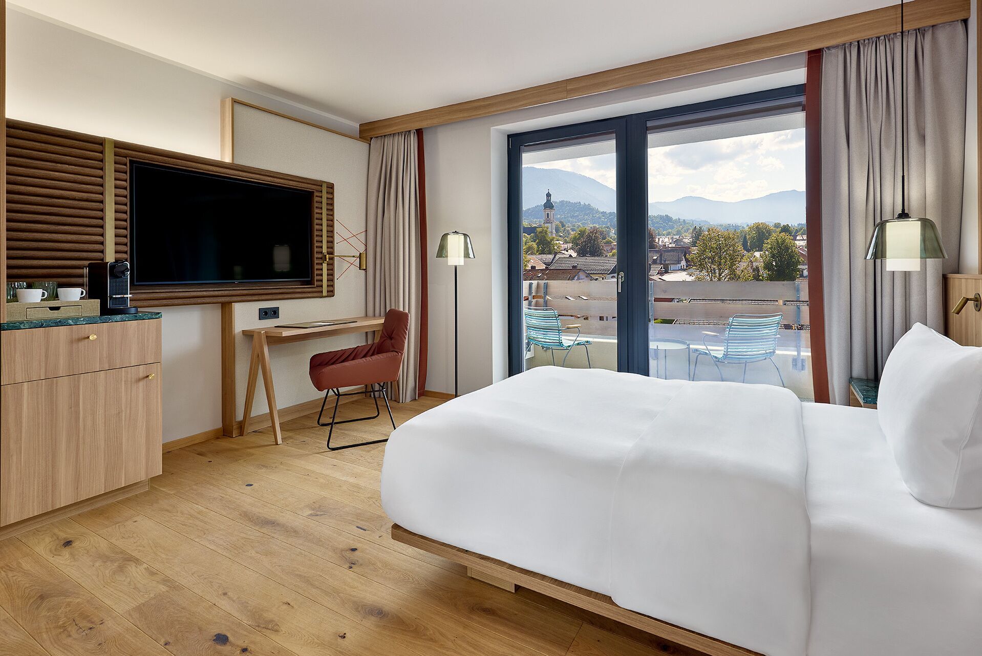 A hotel room with a bed, a desk, a TV, a chair, and a view of the city and mountains.