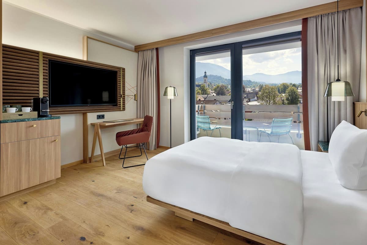 A hotel room with a large bed, a desk with a chair, a television, and a view of the city through a large window.